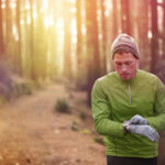 Trail running runner looking at heart rate monitor watch running in forest wearing warm jacket sportswear, hat and gloves. Male jogger running training in woods.