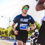 A man running in a fun run. He has sunglasses, a watch, and a race bib