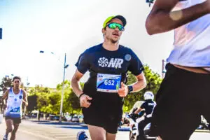 A man running in a fun run. He has sunglasses, a watch, and a race bib
